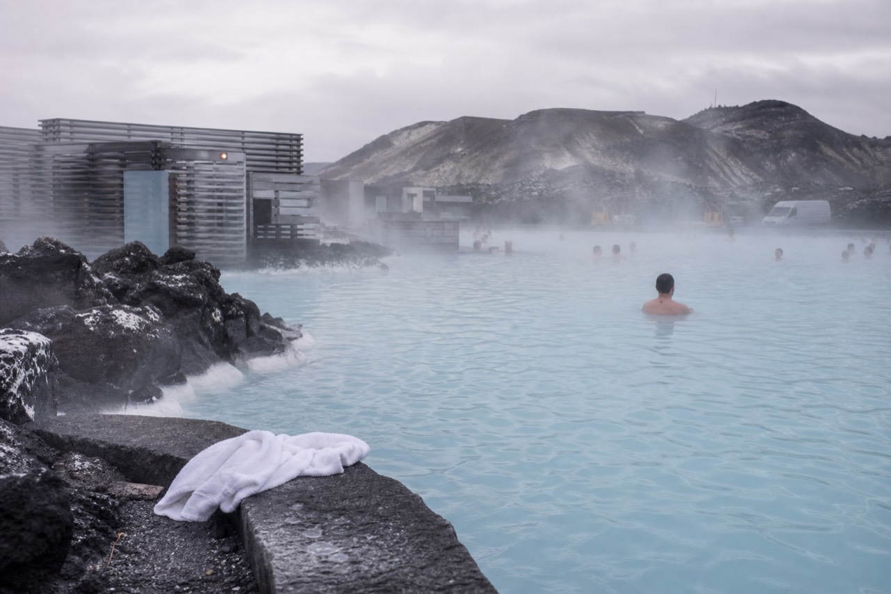 Steam rising over the milky blue water of a geothermal lagoon beside a dark lava field