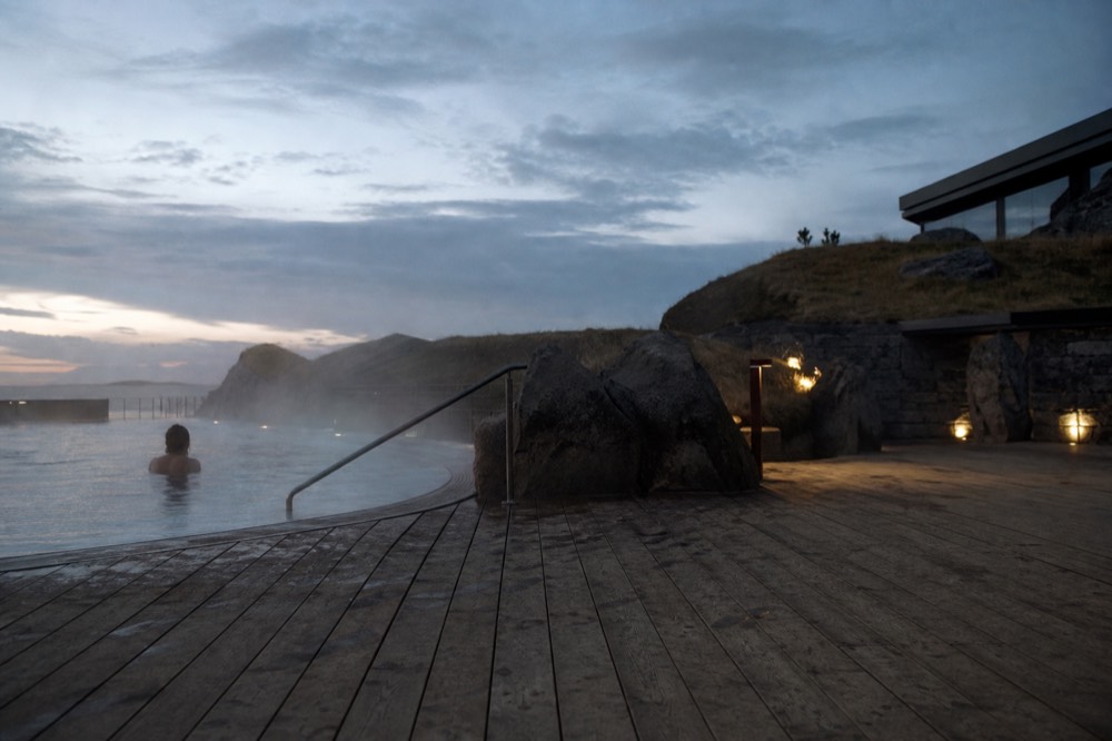 Steam rising off an oceanfront geothermal pool beside a wide wooden deck at dusk