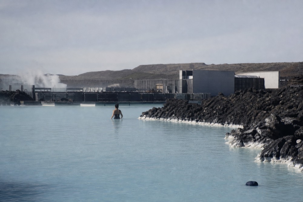 A traveller in a dark bathing suit waist-deep in milky blue water beside a lava ridge