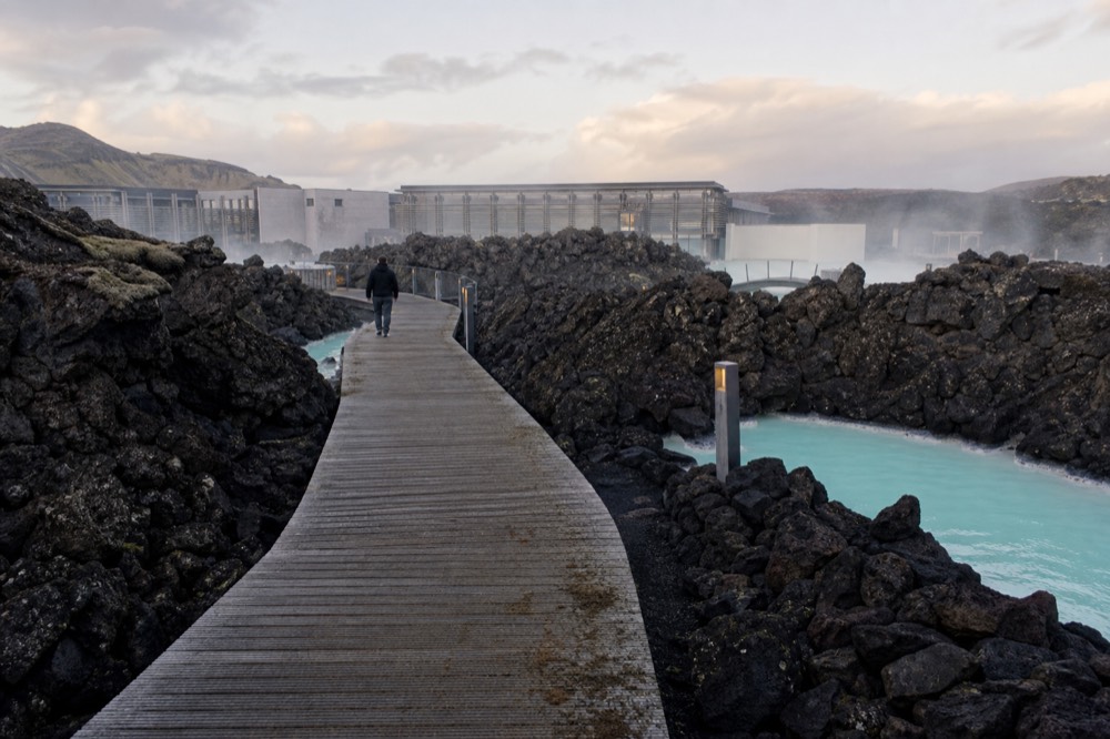 A wooden walkway crossing dark lava fields toward a steaming turquoise lagoon building