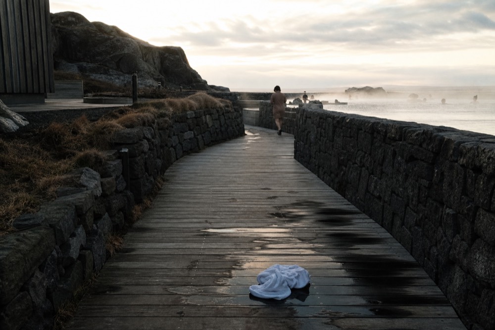 A wooden walkway leading toward a steaming geothermal lagoon in evening light