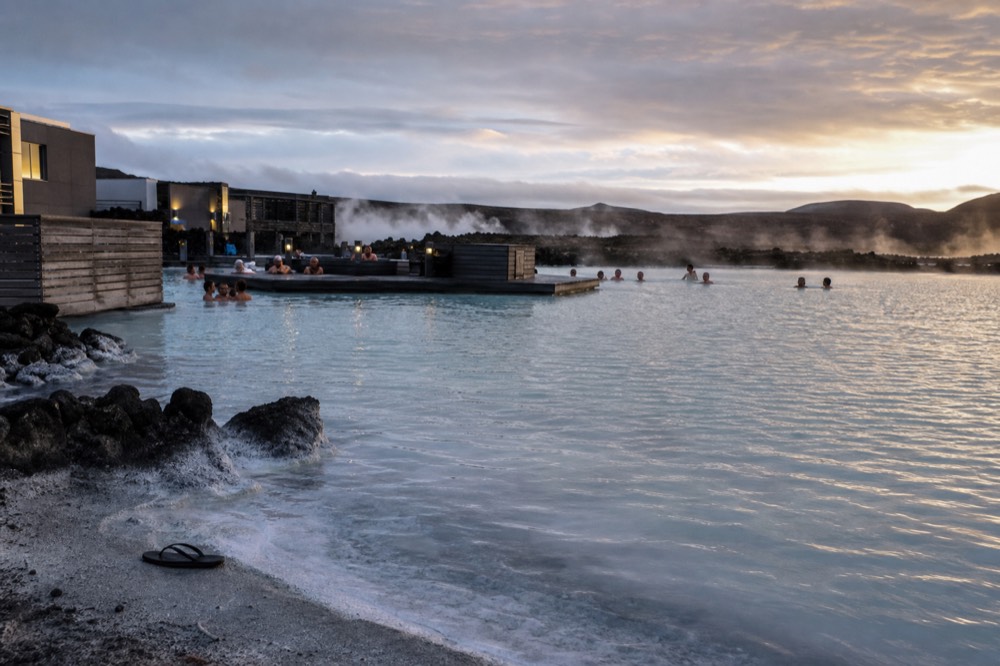 A wide view of a pale blue geothermal lagoon with steam drifting across dark lava fields