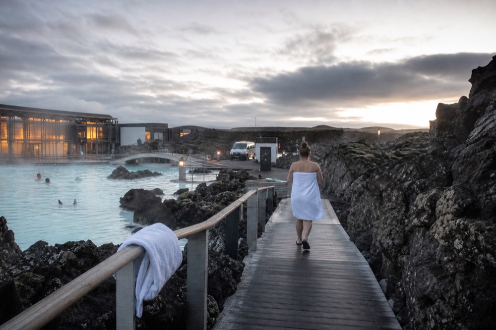A traveller wrapped in a towel walking across a boardwalk beside steaming geothermal water