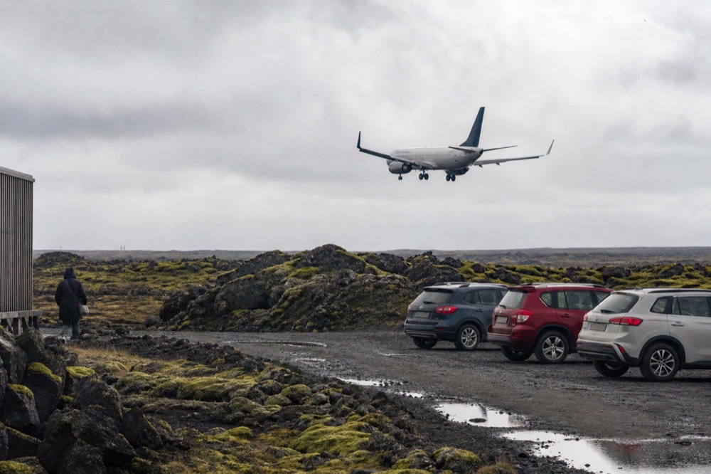 A passenger plane descending low over moss-covered lava fields toward a coastal airport