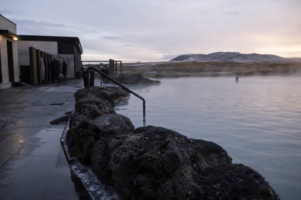 A geothermal lagoon with steam drifting across still water toward distant hills