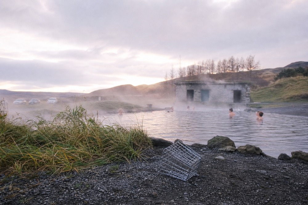 A wide geothermal pool with steam rising from the water and mist over surrounding grass