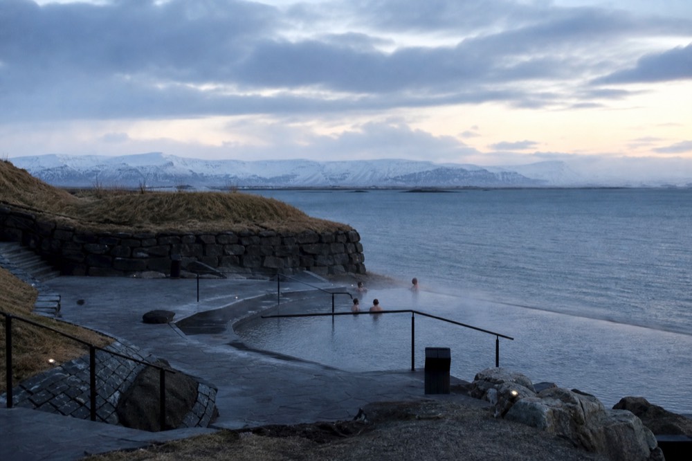 An outdoor geothermal pool on a fjord shoreline with mountains behind and steam over the water