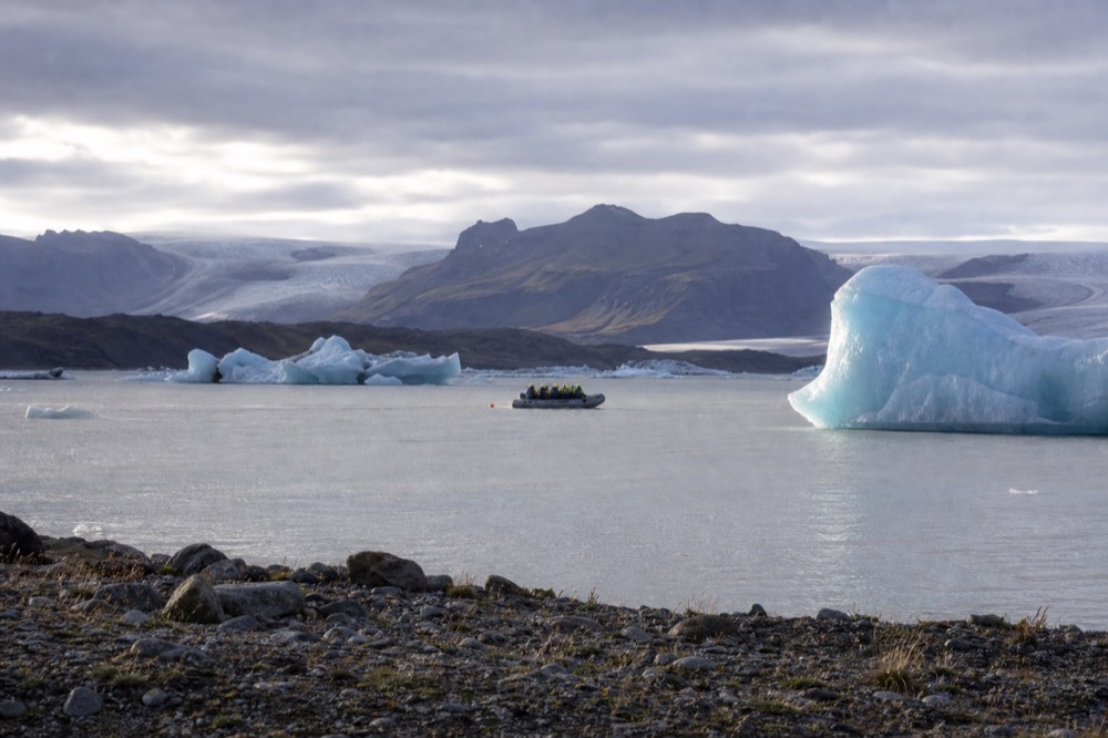 A small boat carrying passengers between large blue icebergs on a grey glacier lagoon