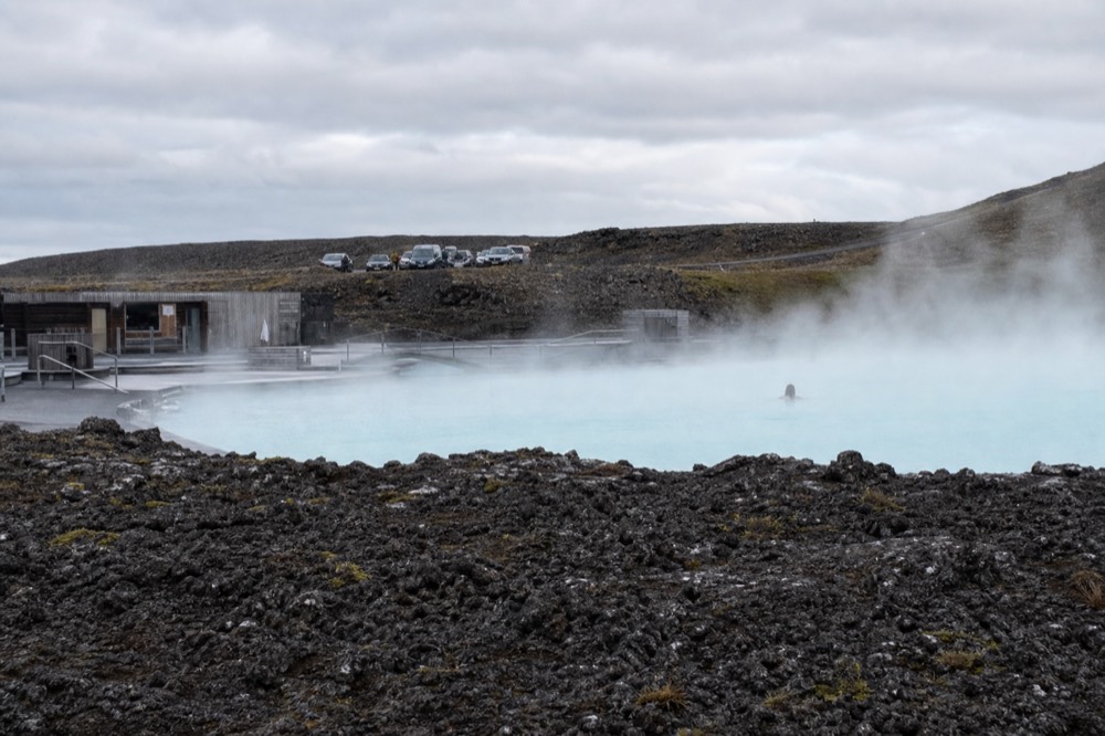 A swimmer's silhouette in steam above a geothermal pool with mountains beyond