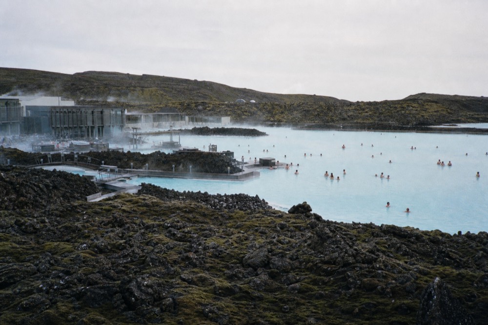 A wide pale blue lagoon steaming gently against dark volcanic rock under a pale sky