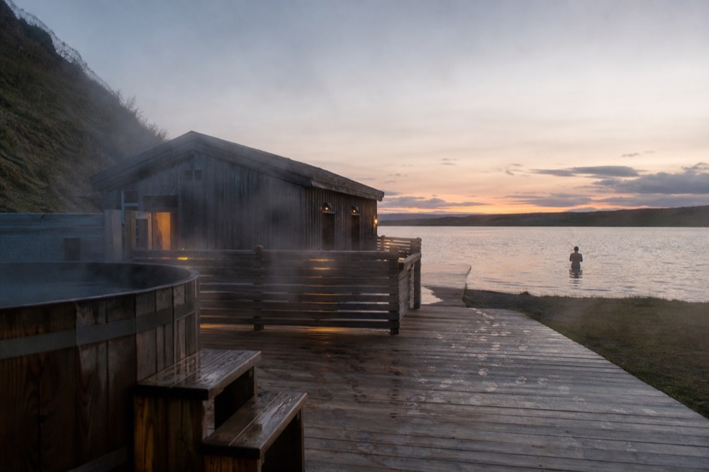 Geothermal steam rising from a modern bathhouse beside a calm lake at dusk