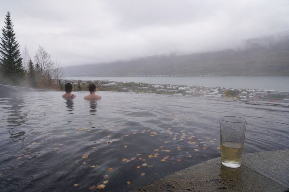 Steam rising from a hot pool on a forested slope with a town and fjord visible below