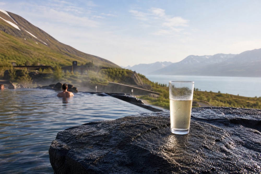 Hands resting a drink glass on the stone edge of a hot pool with steam drifting past