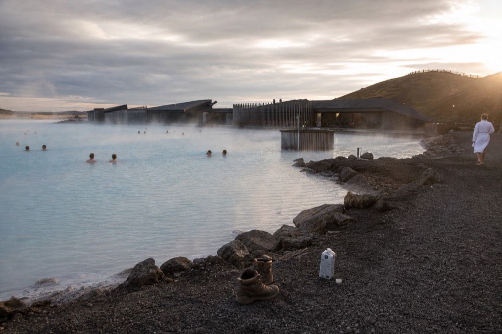 A geothermal pool beside a dark lava field with steam rising in low evening light