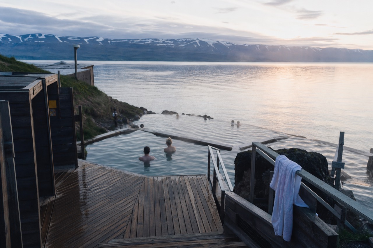 Steaming geothermal pools on a clifftop overlooking a wide bay with low hills on the far shore