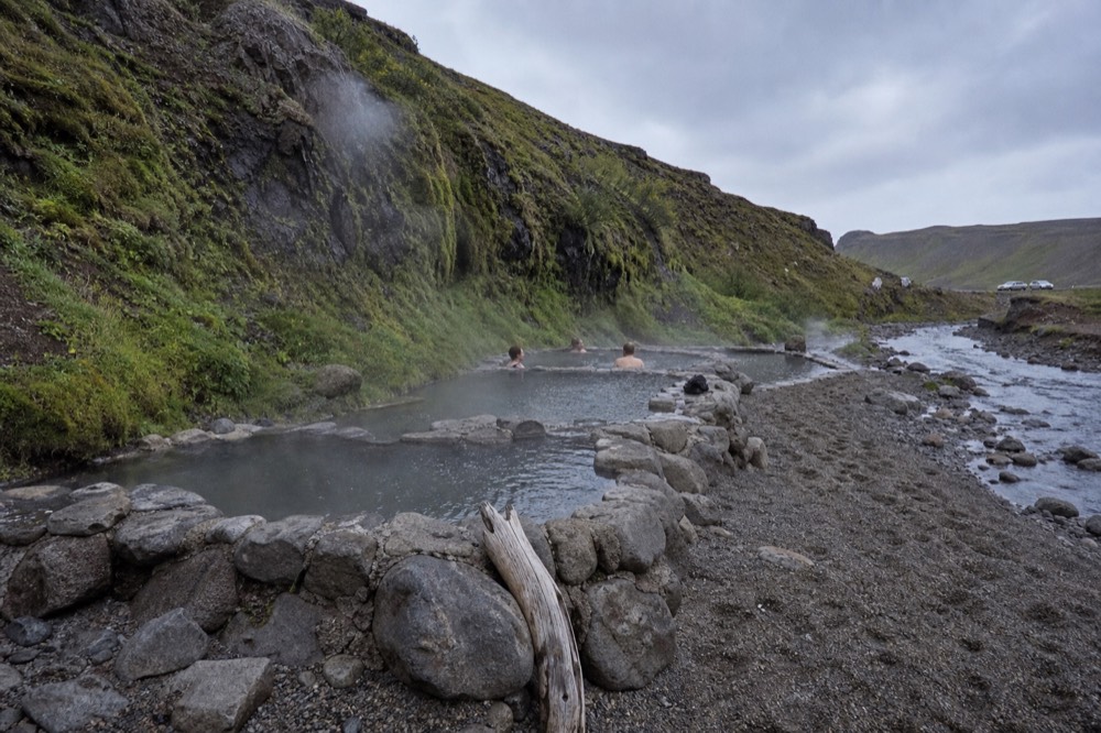 Three linked geothermal pools with steam rising beside a gravel plain and dark mountain ridges