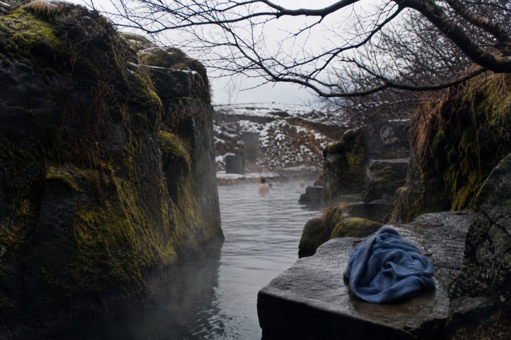 A narrow canyon in West Iceland with steam rising from still water between dark rock walls
