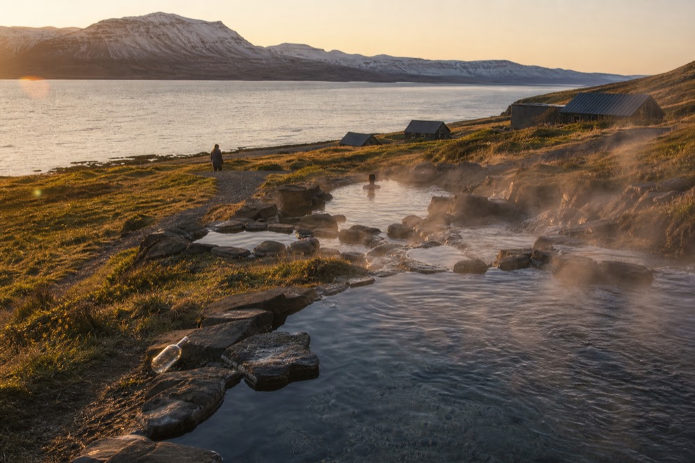 Steaming pools terraced down a grassy slope toward a calm ocean fjord