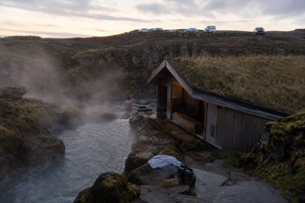 A wooden bathhouse with a turf roof beside a steaming pool in a rocky gorge
