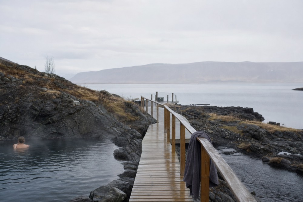 A wooden walkway between steaming pools with fjord water beyond