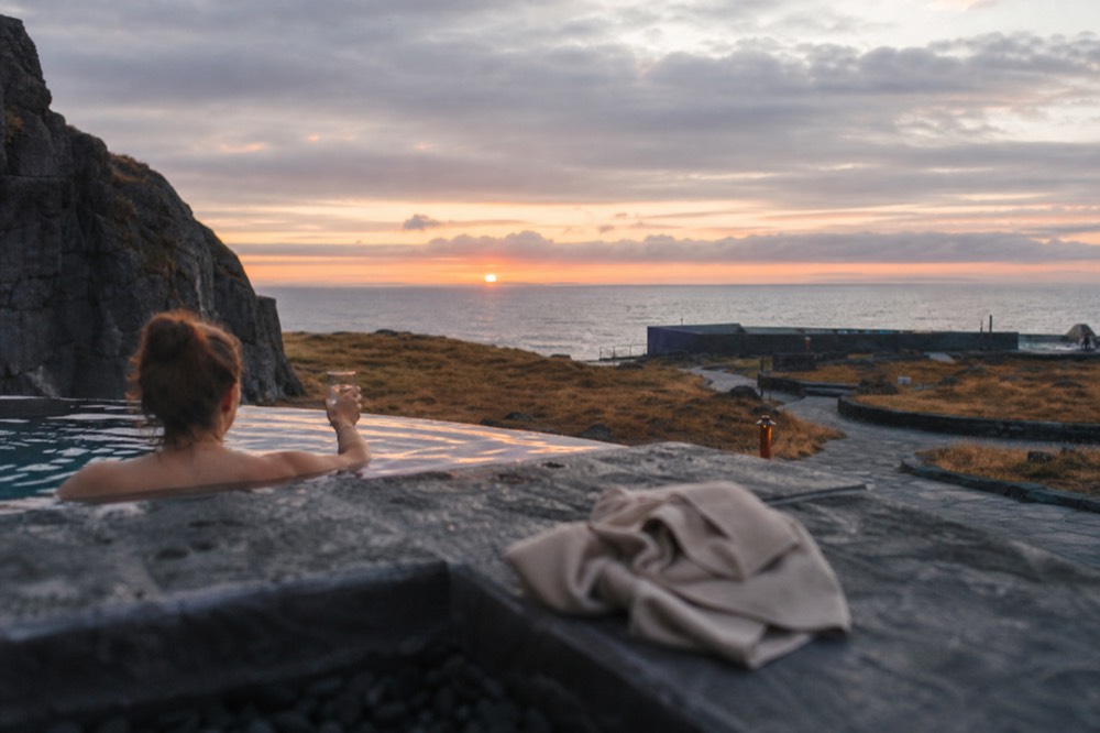 A lagoon visitor holding a drink while soaking in a geothermal pool with a towel visible on the pool edge