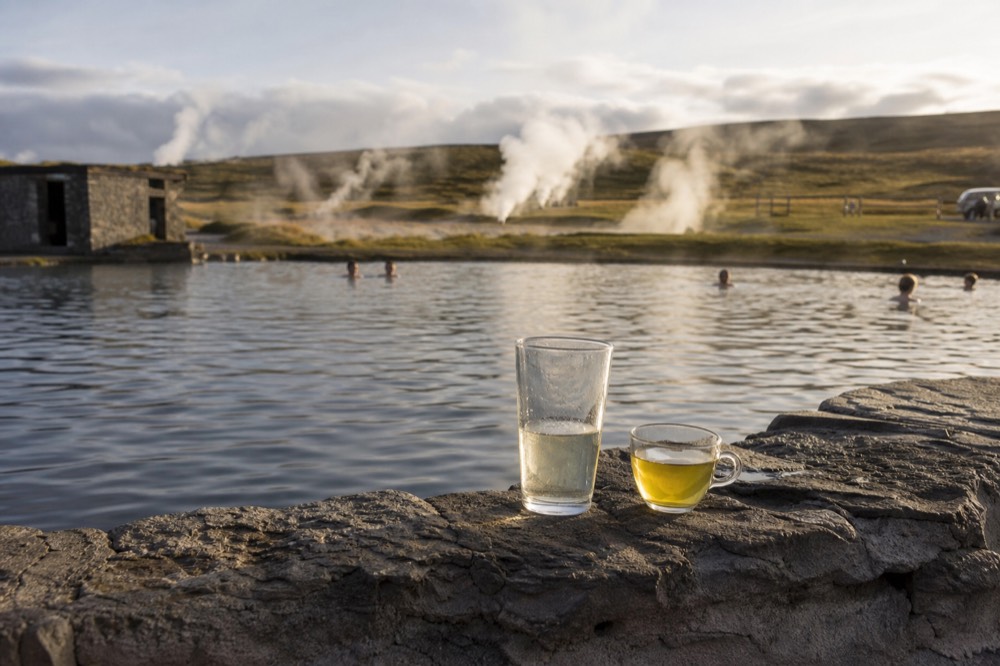 A tall glass and a small herbal tea cup on a stone ledge beside steaming pool water