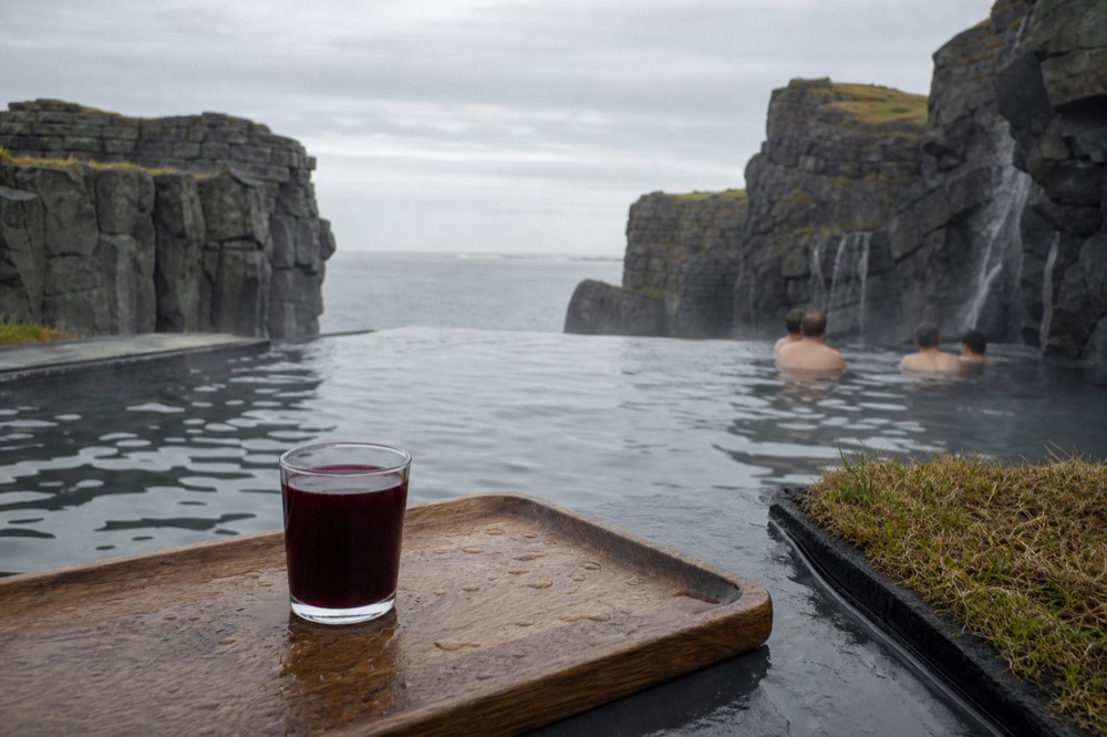 A small glass of dark berry juice on a wooden tray beside a steaming infinity pool at the ocean's edge