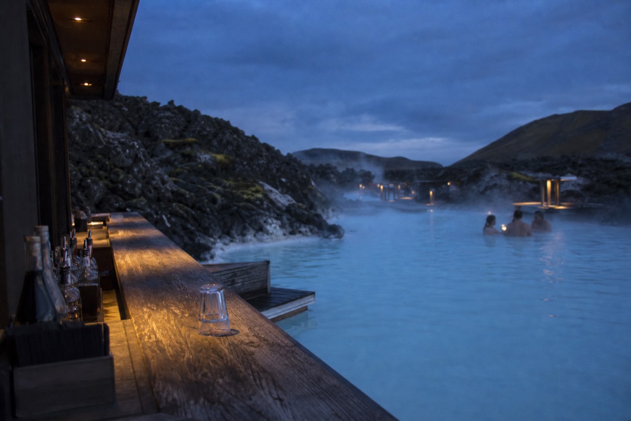 Steam rising from a blue geothermal lagoon beside a wooden bar counter at dusk