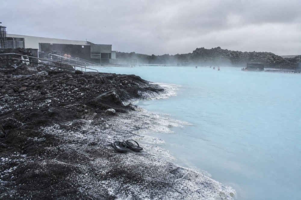A pale blue geothermal lagoon spreading across a dark lava field under an overcast sky