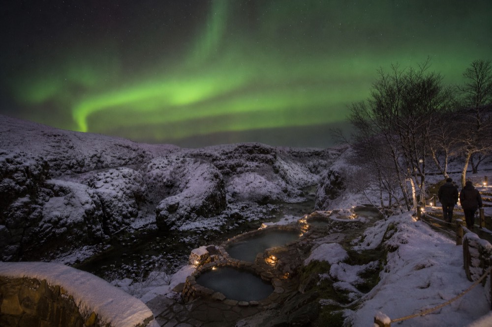 Green aurora curtains over a snowy landscape with a faint glow of steam in the foreground