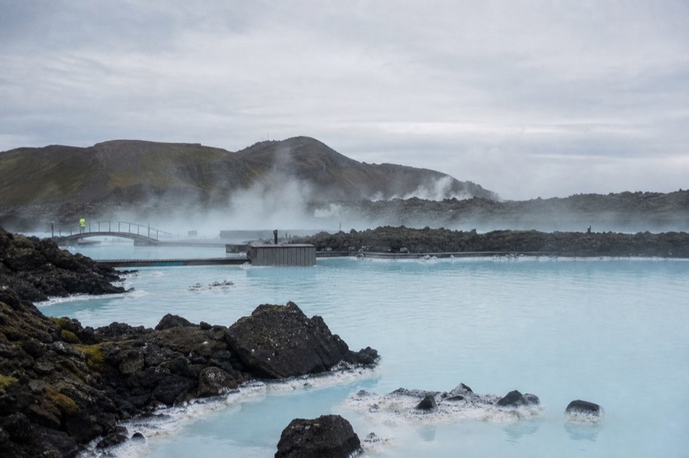 A milky blue geothermal pool spreading wide across a dark lava field under an overcast sky