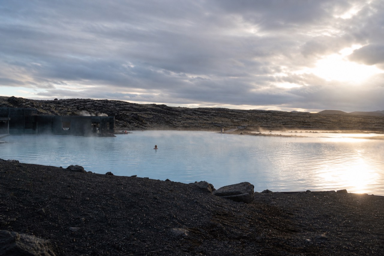 Steam rising from a geothermal lagoon beside a dark volcanic landscape under a wide sky