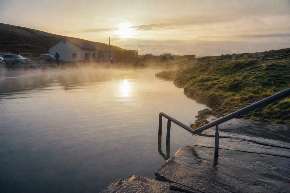 A calm geothermal pool with steam drifting across still water in evening light