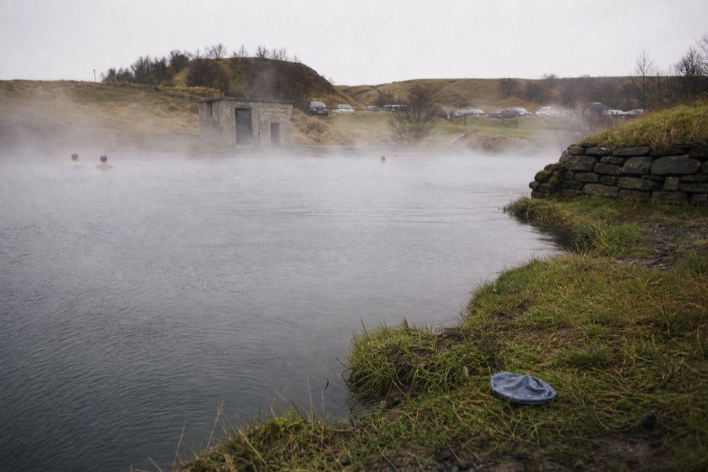 A natural geothermal pool with steam drifting across still water and grassy banks