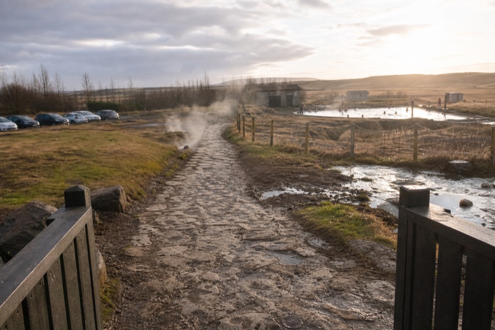 A gravel path leading past small steaming hot springs toward a geothermal pool
