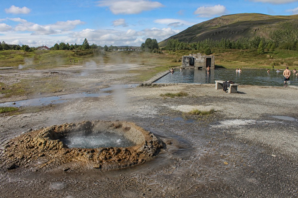 An open geothermal field with steam rising from several small springs beside a still pool