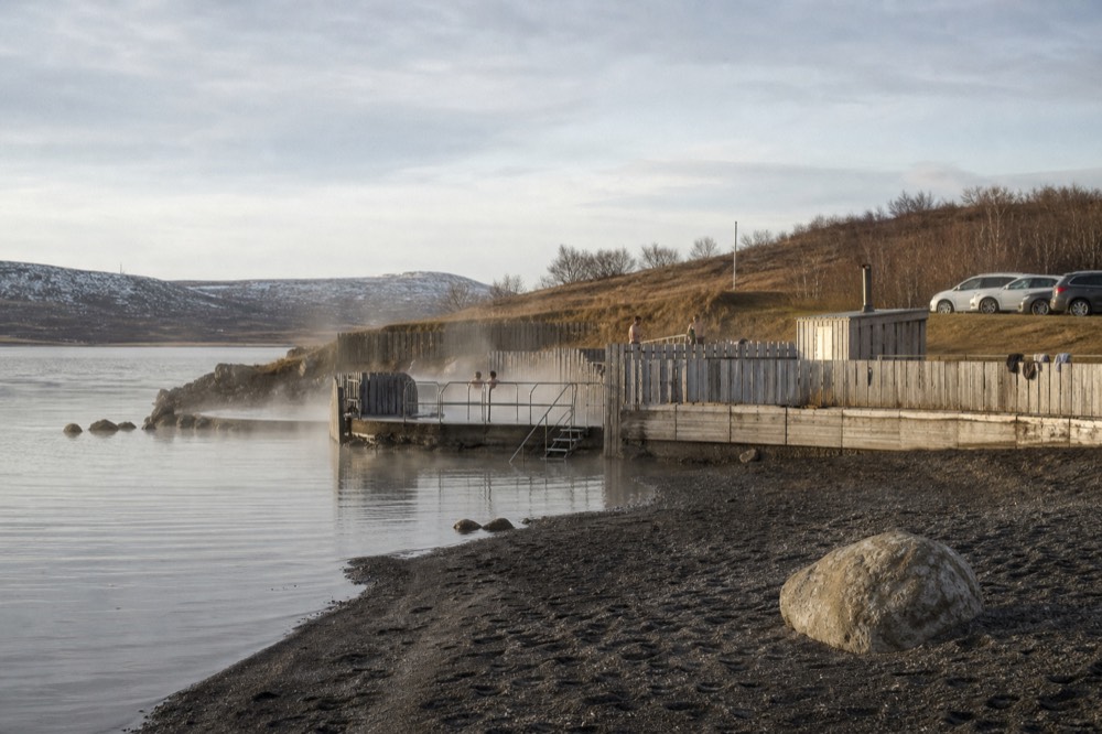 A lakeside geothermal bathing complex with steam rising over still water