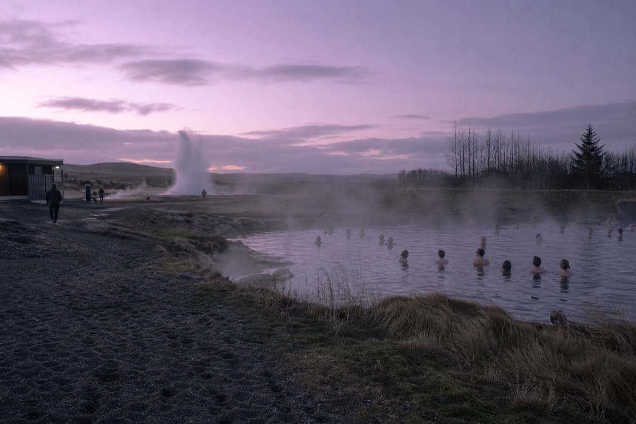 Steam rising from a geothermal pool surrounded by grass and small hot springs at dusk
