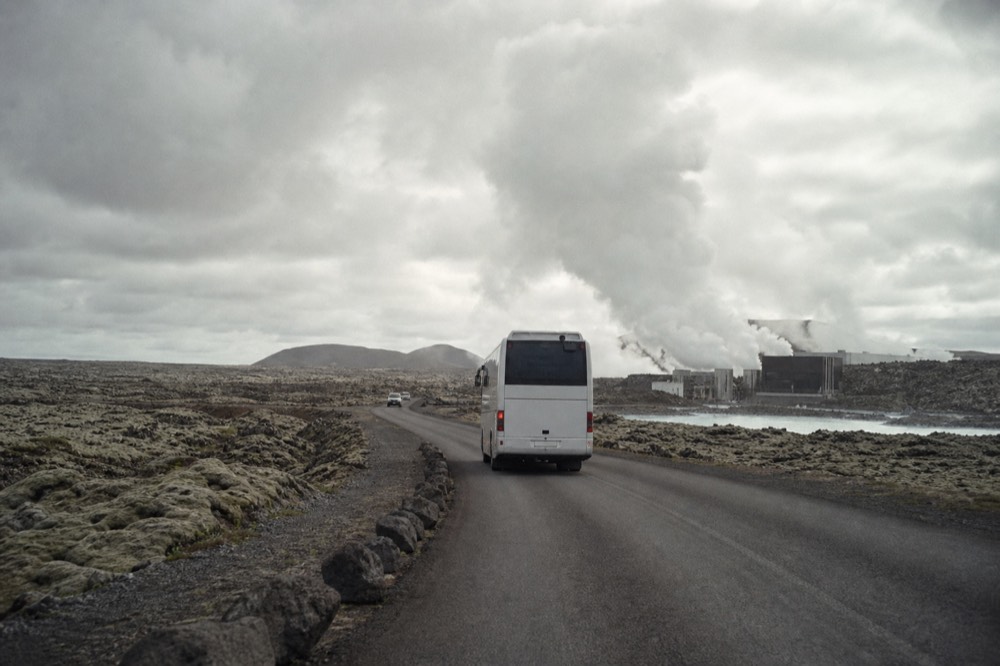 A coach on a coastal road approaching a steam plume on a lava plain