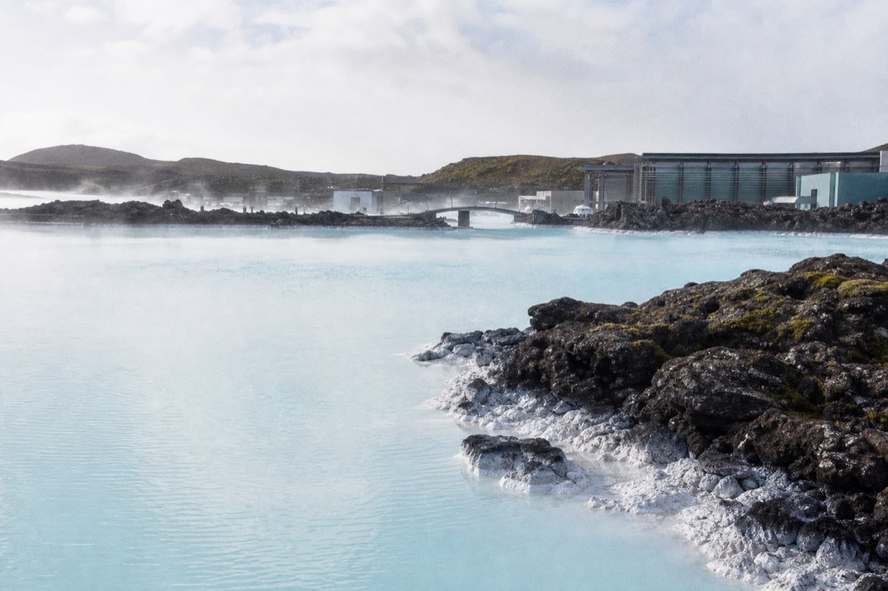 Steam rising from a still blue geothermal lagoon surrounded by dark lava rock under a pale sky